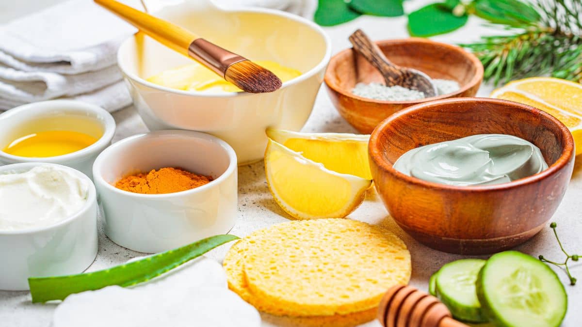 Various natural skincare ingredients arranged on a light surface, including lemon slices, cucumbers, honey, and bowls with creams and powders.