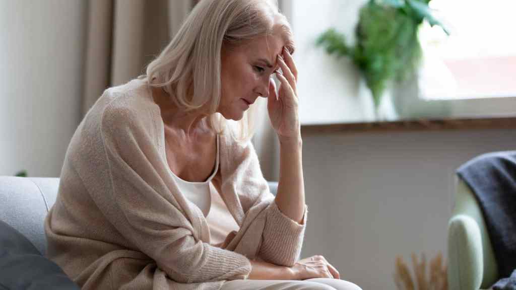 Older woman with light hair sits on a couch indoors, resting her head on her hand and looking down with a concerned expression.