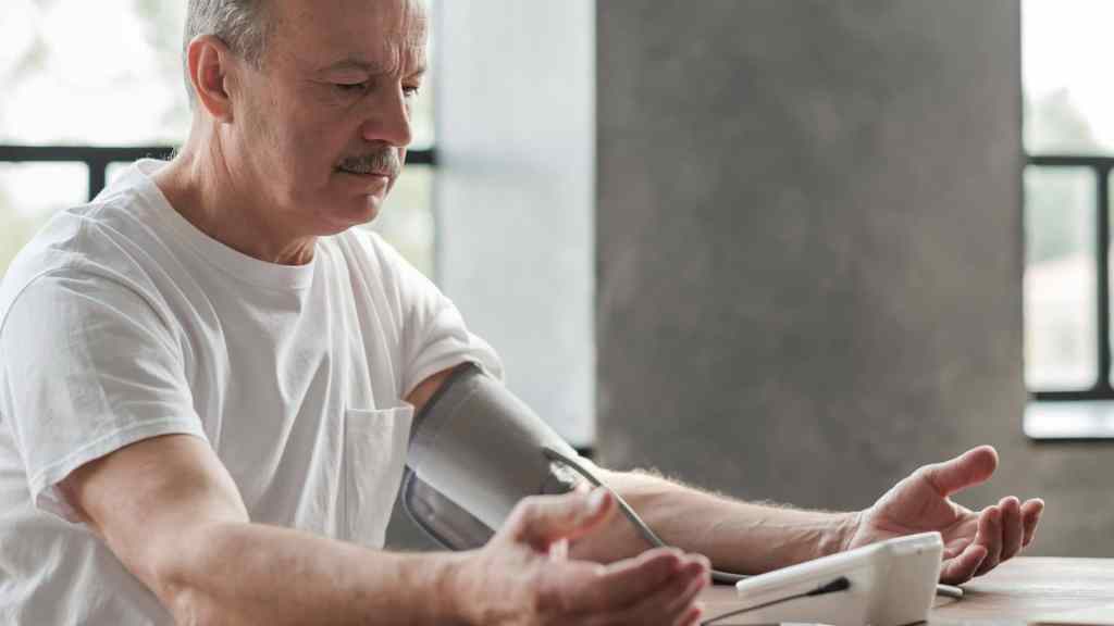 Older man in a white t-shirt sits at a table checking his blood pressure with an electronic monitor, looking at the device's display.
