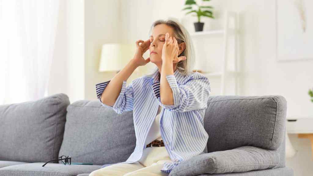 An older woman sits on a couch with her eyes closed, pressing her fingers to her temples. Glasses rest nearby on the sofa. The background shows a bright, neatly decorated living room.