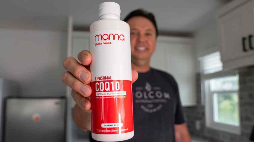 A person holds a bottle labeled “Manna Liposomal CoQ10” with a red and white design in a kitchen setting.