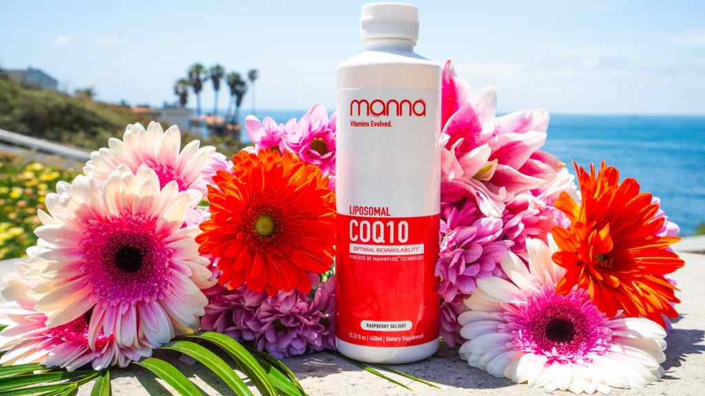 A bottle of Manna Liposomal CoQ10 supplement is placed on a stone surface, surrounded by colorful flowers, with the ocean and palm trees in the background.