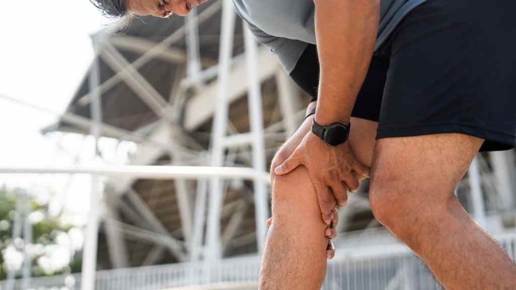 A person outdoors in athletic wear bends over and holds their knee, appearing to be in pain or discomfort.