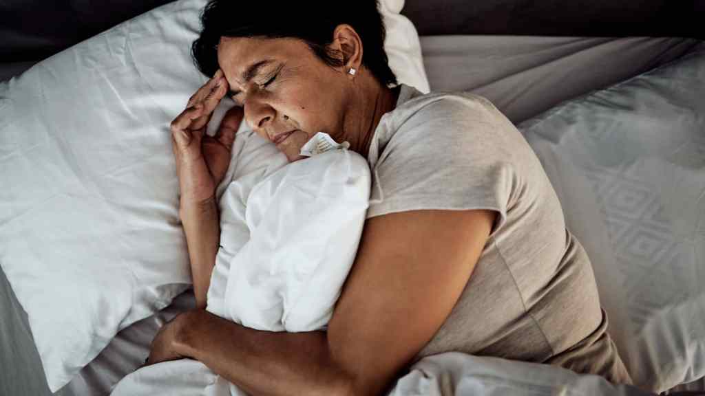 Woman lying in bed holding her head and appearing to be in discomfort or pain, with her eyes closed and hand on her forehead.