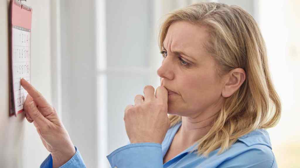 A woman in a blue shirt looks at a wall calendar, appearing thoughtful and concerned, with her finger on her lips.