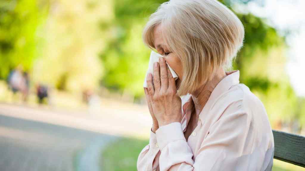 An older woman sitting on a bench outdoors wipes or blows her nose with a tissue. Trees and a path are visible in the blurred background.