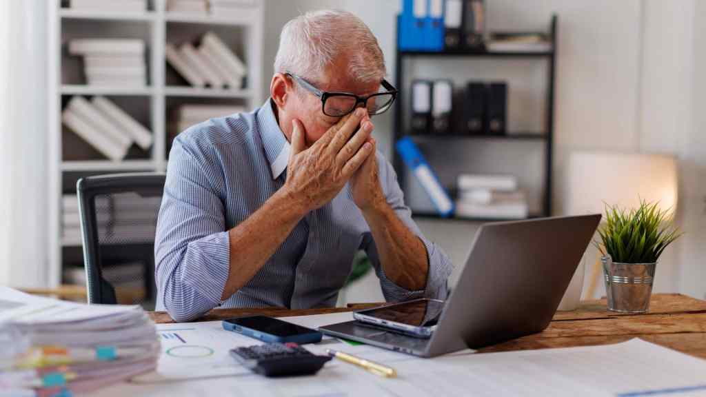 An older man sits at a desk with a laptop, covering his face with his hands, appearing stressed or tired in an office setting.