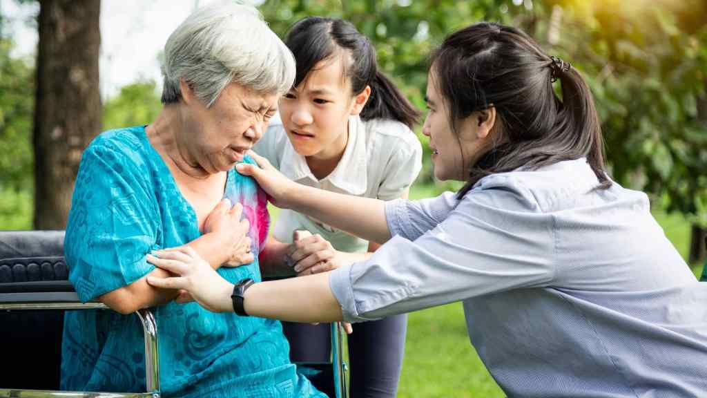 An elderly woman in a wheelchair clutches her chest in pain while two younger women support and assist her outdoors.