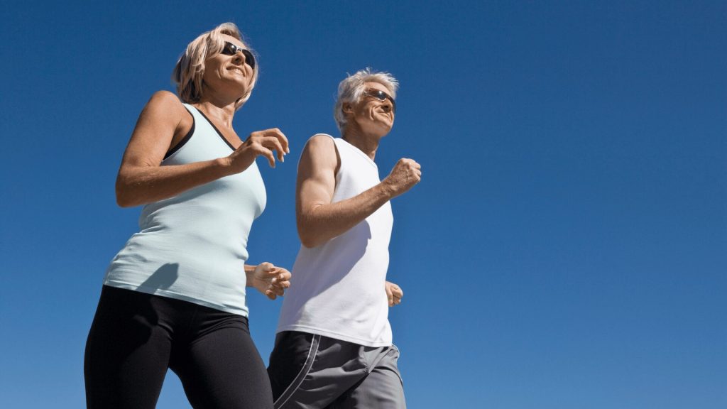 Two older adults in athletic clothing are walking outdoors under a clear blue sky. They are smiling and wearing sunglasses.