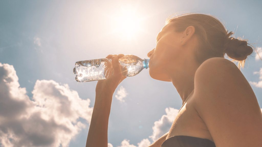 A person with hair tied back drinks water from a plastic bottle while standing outdoors under a bright sun and blue sky with clouds.
