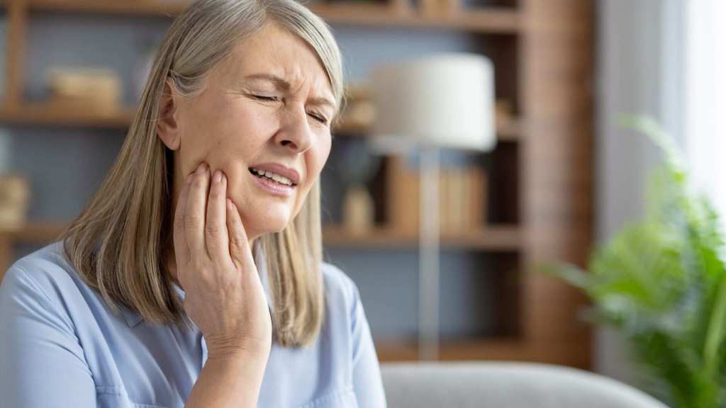 An older woman sits indoors, eyes closed, touching her jaw with a painful expression, possibly experiencing tooth or jaw pain.