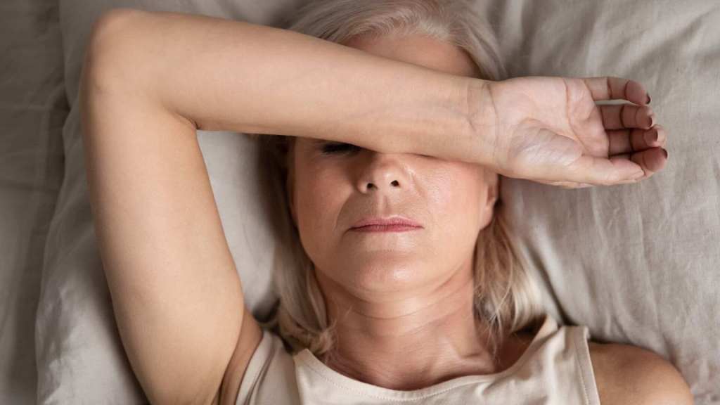A woman with light hair lies on a bed, covering her eyes with her arm, appearing tired or stressed.