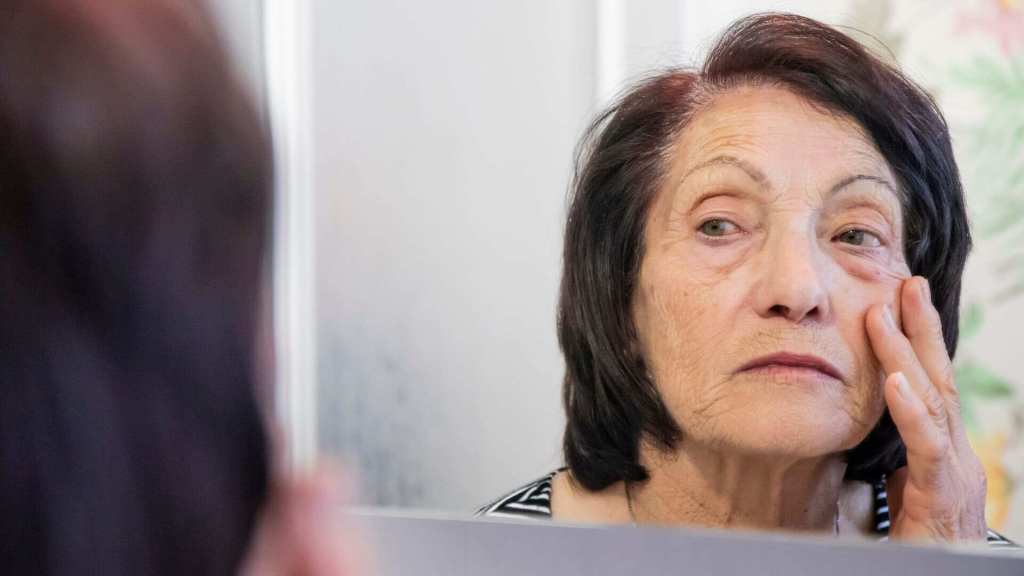 An older woman with dark hair looks at her reflection in a mirror while gently touching the skin below her eye.