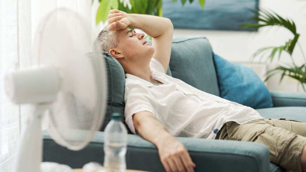 A man in light clothing sits on a couch, looking overheated and tired, with a fan and a water bottle nearby in a bright living room.