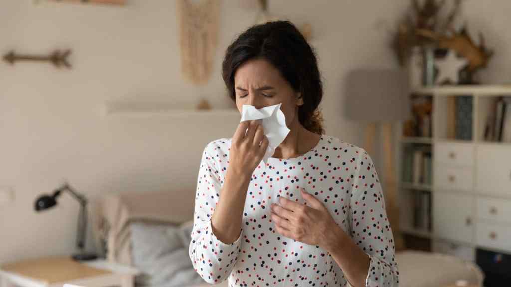 A person indoors holds a tissue to their nose and closes their eyes, appearing to sneeze or have a cold.