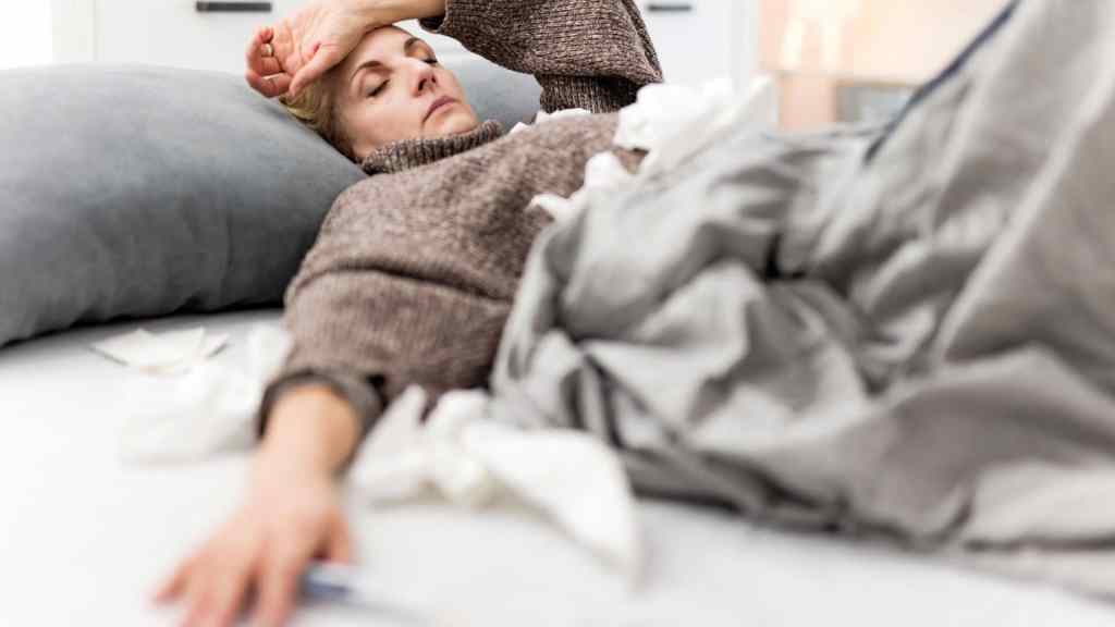 Woman lying on a bed looking unwell, covered with a blanket, surrounded by tissues, with one arm extended and a thermometer nearby.