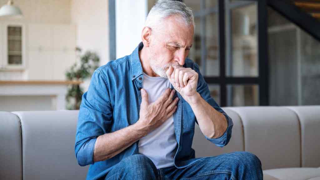 Older man sitting on a couch, coughing into his hand and holding his chest, appearing to experience discomfort or respiratory symptoms.