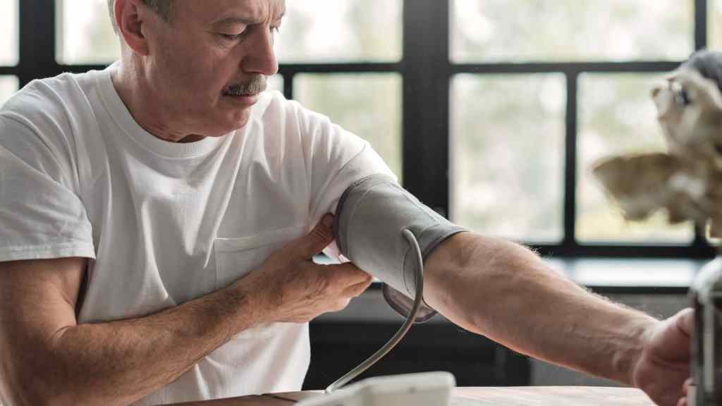An older man in a white shirt sits indoors and measures his blood pressure with an electronic monitor.