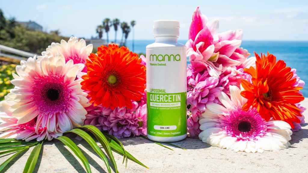 A bottle of Mamá Liposomal Quercetin sits on a stone surface surrounded by colorful gerbera daisies and palm leaves, with the ocean and palm trees in the background.