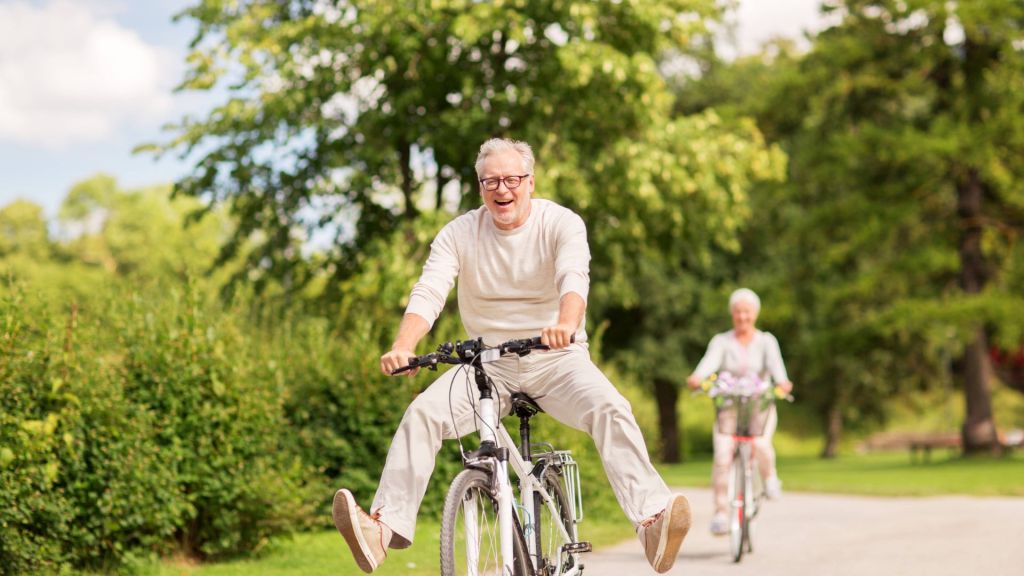An elderly man is riding a bicycle with his legs outstretched and smiling, followed by an elderly woman on a bicycle. They are on a path in a park with trees and greenery in the background.