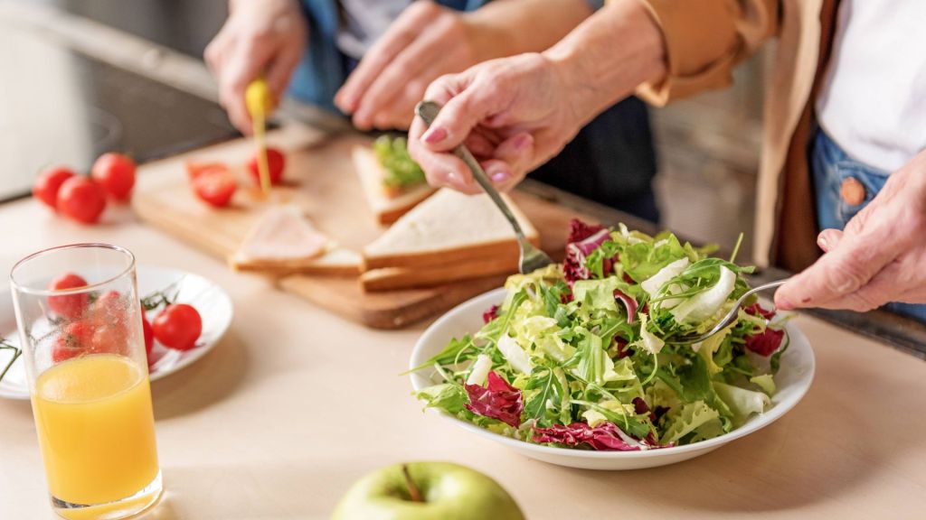 Two people preparing a meal with one person making salad in a bowl and the other slicing tomatoes and sandwiches on a cutting board. A glass of orange juice and an apple are on the counter.