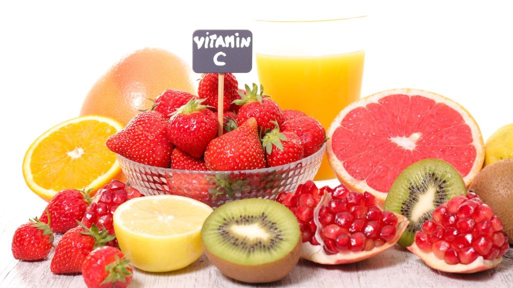 A variety of fruits high in vitamin C, including strawberries, kiwi, grapefruit, pomegranate, and lemon, displayed with a glass of orange juice. A small sign indicates "Vitamin C.