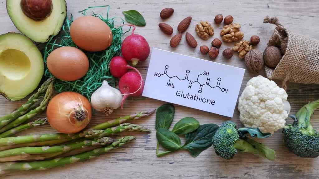 Various foods like avocados, eggs, nuts, radishes, cauliflower, broccoli, asparagus, onions, garlic, and spinach are arranged around a card with a glutathione chemical structure.