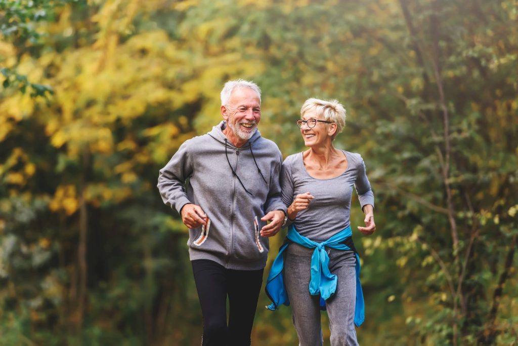 An older man and woman are jogging outdoors on a path with lush green foliage in the background, both smiling and dressed in casual exercise clothing.