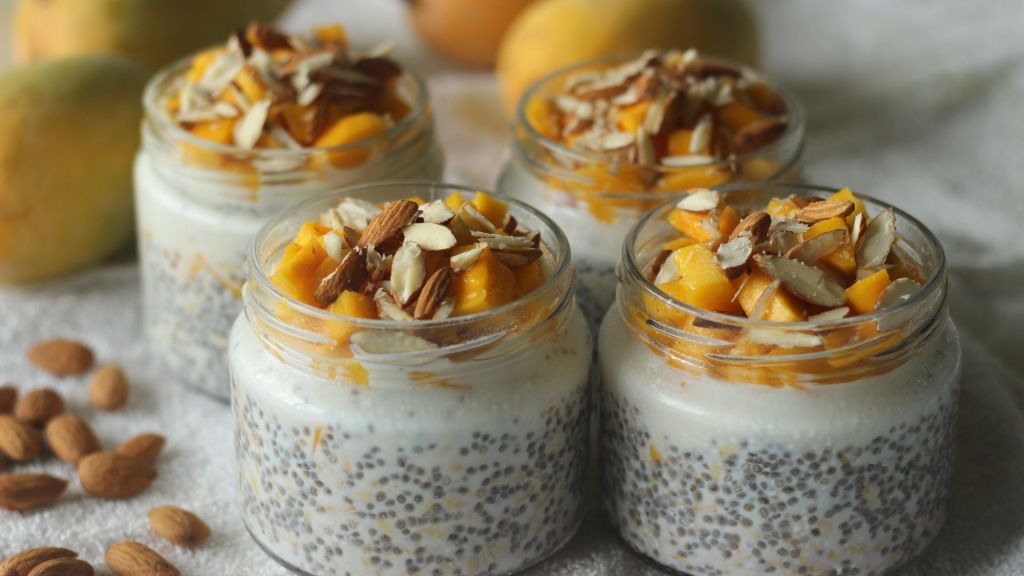 Four jars of chia pudding topped with mango chunks and sliced almonds, placed on a cloth with scattered almonds nearby.