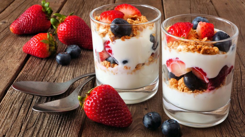 Two glasses of yogurt parfait with strawberries, blueberries, and granola on a wooden table, accompanied by two spoons.
