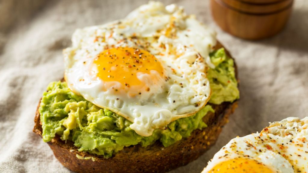Avocado toast topped with a sunny-side-up egg, sprinkled with pepper, on a slice of whole-grain bread.