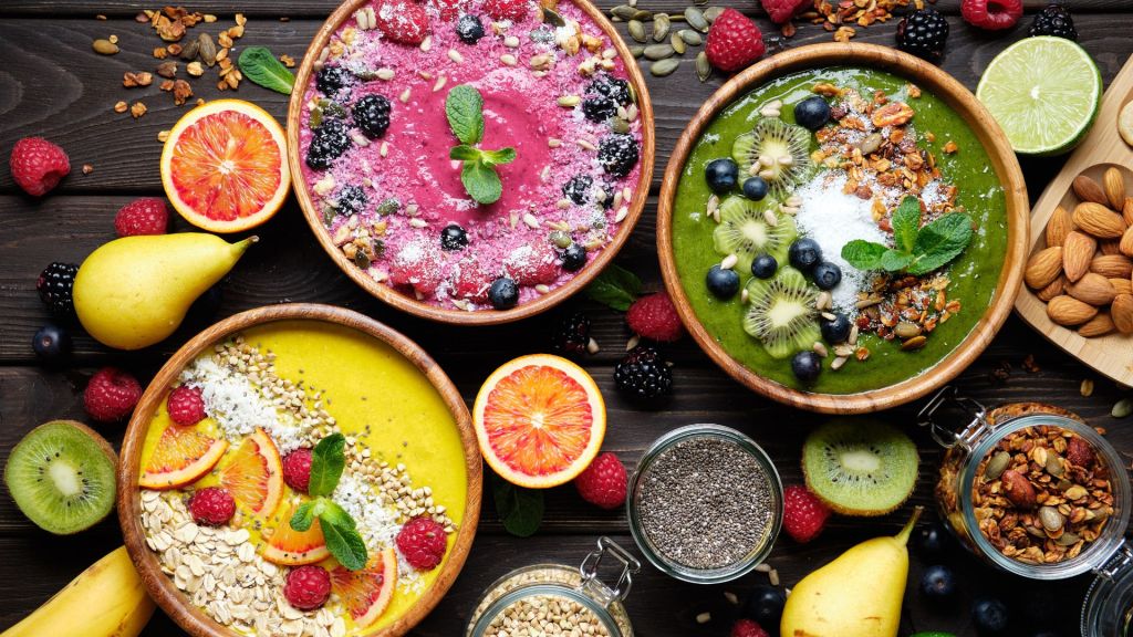 Three smoothie bowls with assorted toppings including berries, kiwis, mint leaves, seeds, and nuts, surrounded by pears, raspberries, a lime, and various seeds and nuts on a dark wooden table.