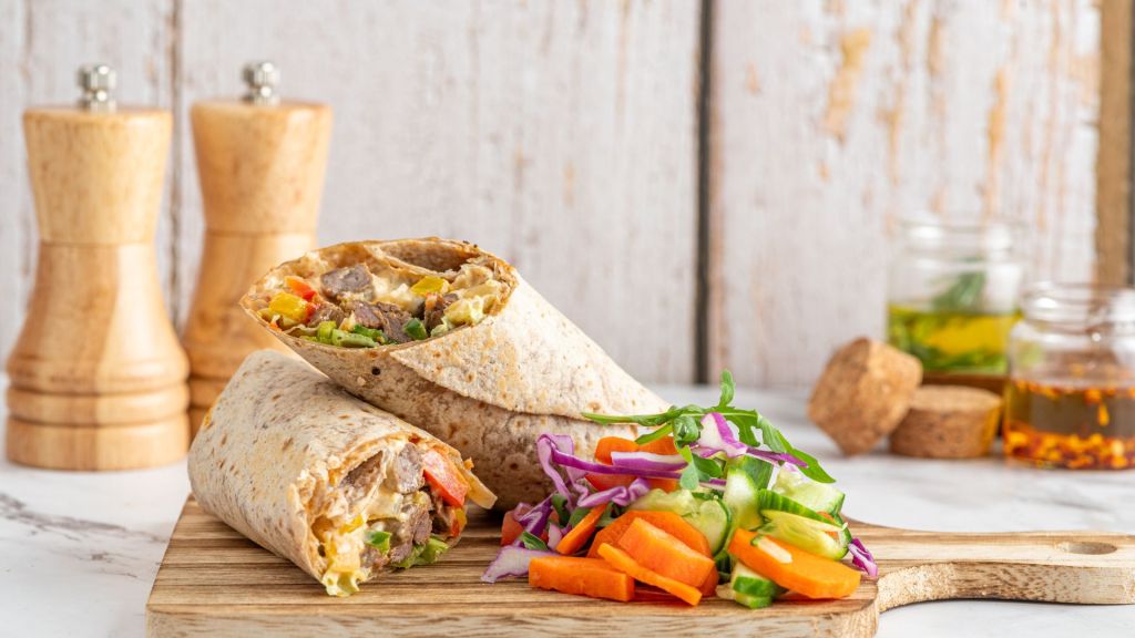 Two burritos filled with meat and vegetables on a wooden board, served with sliced carrots, cucumber, and purple cabbage. Pepper mills and jars in the background.