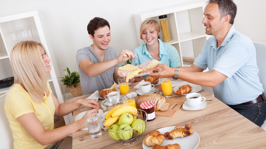 Four people enjoy breakfast together, sharing bread, fruit, and pastries at a wooden table.