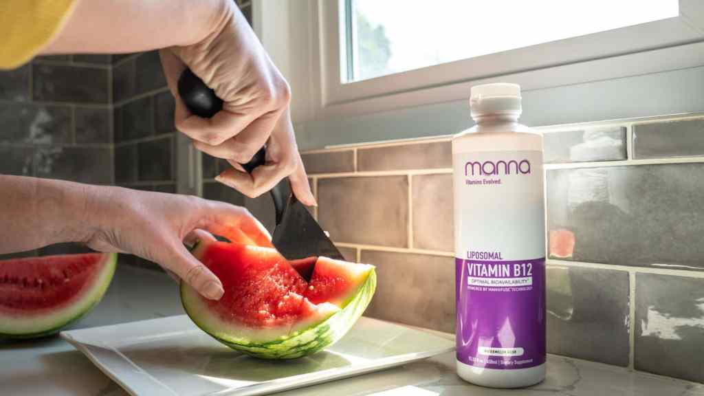 A person slices a watermelon on a kitchen counter next to a bottle labeled “Liposomal Vitamin B12” by the brand Manna.