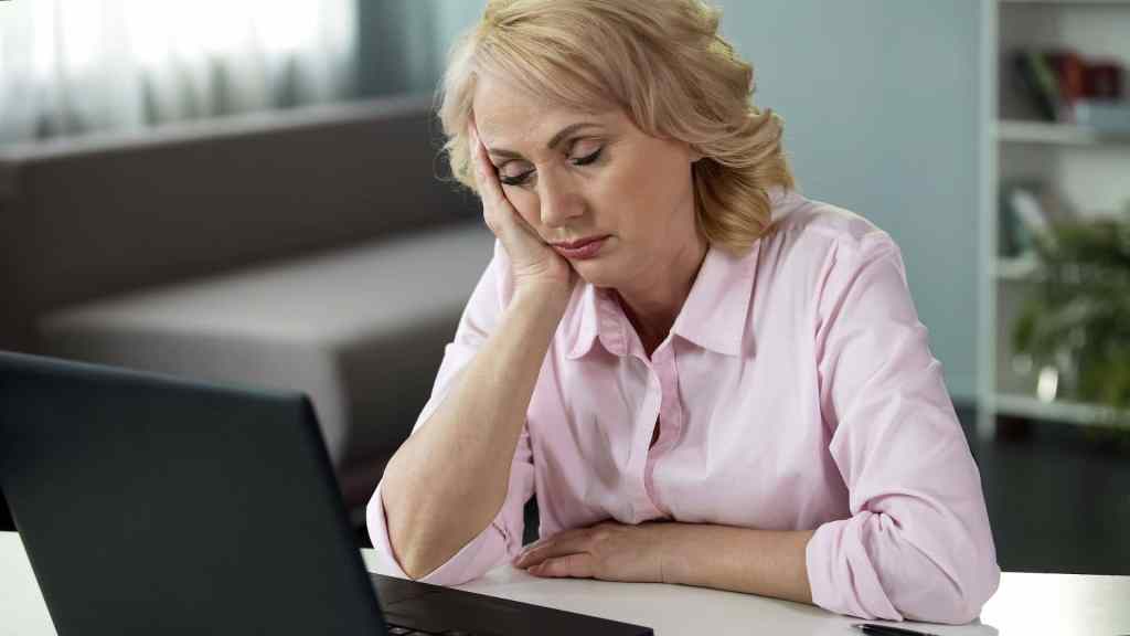 A woman with blond hair wearing a pink shirt sits at a table, resting her head on her hand and looking tiredly at a laptop screen.