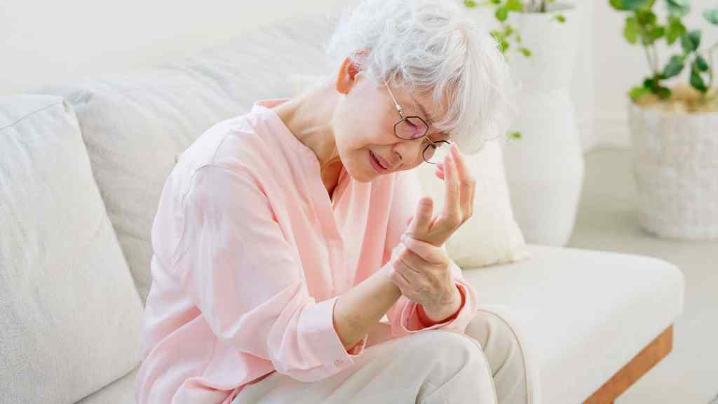 Older woman with short white hair sitting on a sofa, holding her wrist and appearing to be in pain, with houseplants visible in the background.