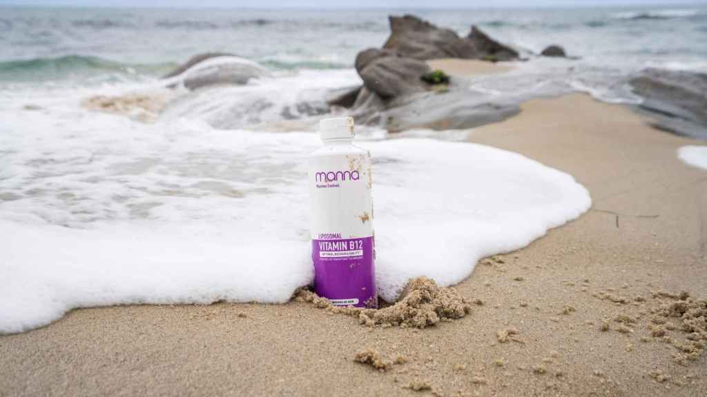 A vitamin B12 drink bottle partially buried in sand near waves on a beach.