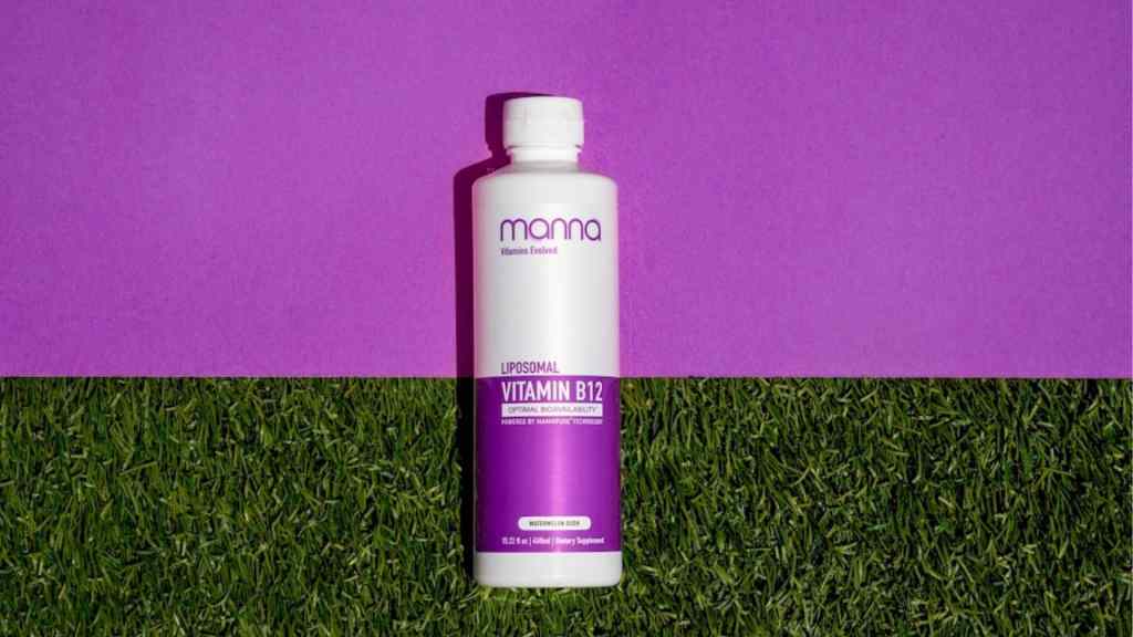 A bottle of Mana Liposomal Vitamin B12 dietary supplement with a purple label rests on artificial grass against a purple background.