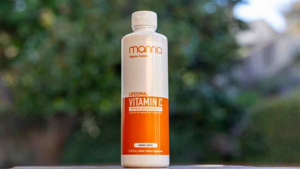 A bottle of Manna Liposomal Vitamin C with an orange gradient label stands against a blurred green and grey outdoor background.