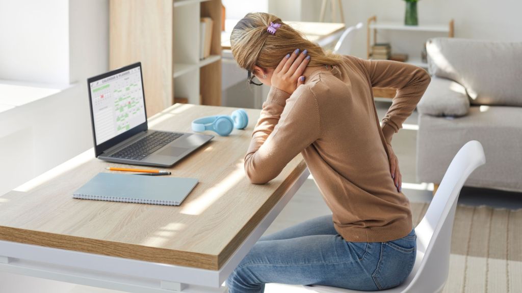 Woman experiencing neck pain while working on a laptop at a home office desk.