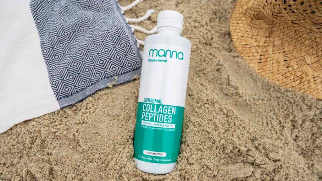 A bottle of liposomal collagen peptides supplement rests on sandy ground next to a patterned cloth and a woven hat.