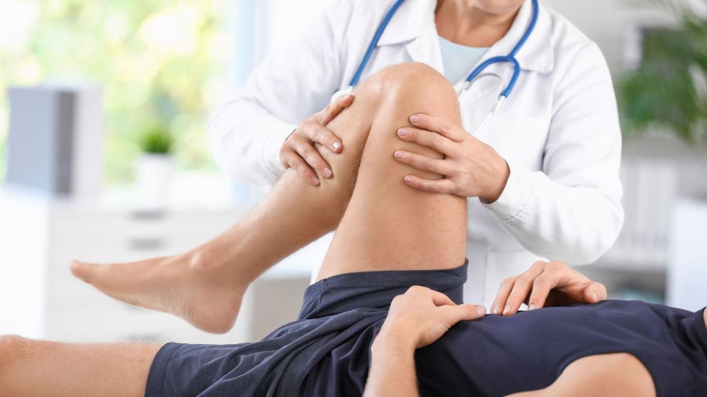 A medical professional wearing a white coat and stethoscope examines and manipulates the knee of a patient lying down.