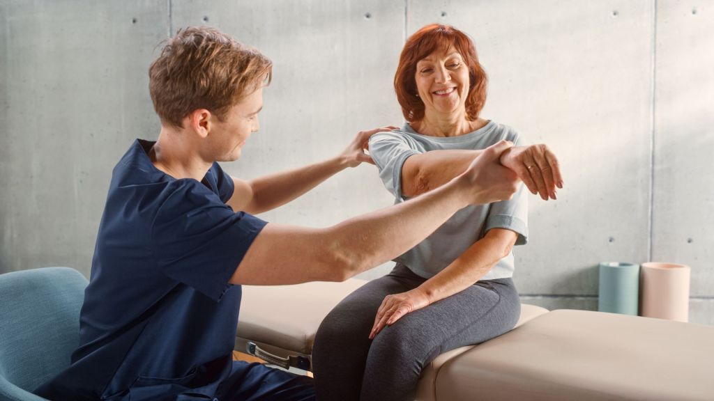 A physical therapist assists a woman in performing arm stretches on an examination table in a clinic.
