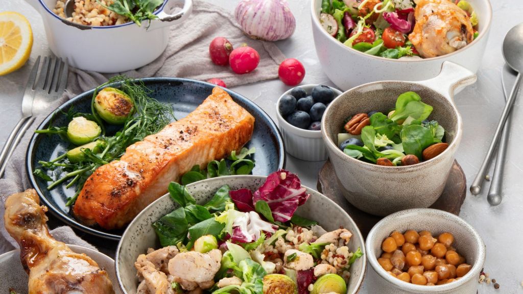 A table with various dishes including salmon, chicken drumstick, salads, berries, chickpeas, and brussels sprouts. Utensils are placed around the dishes.