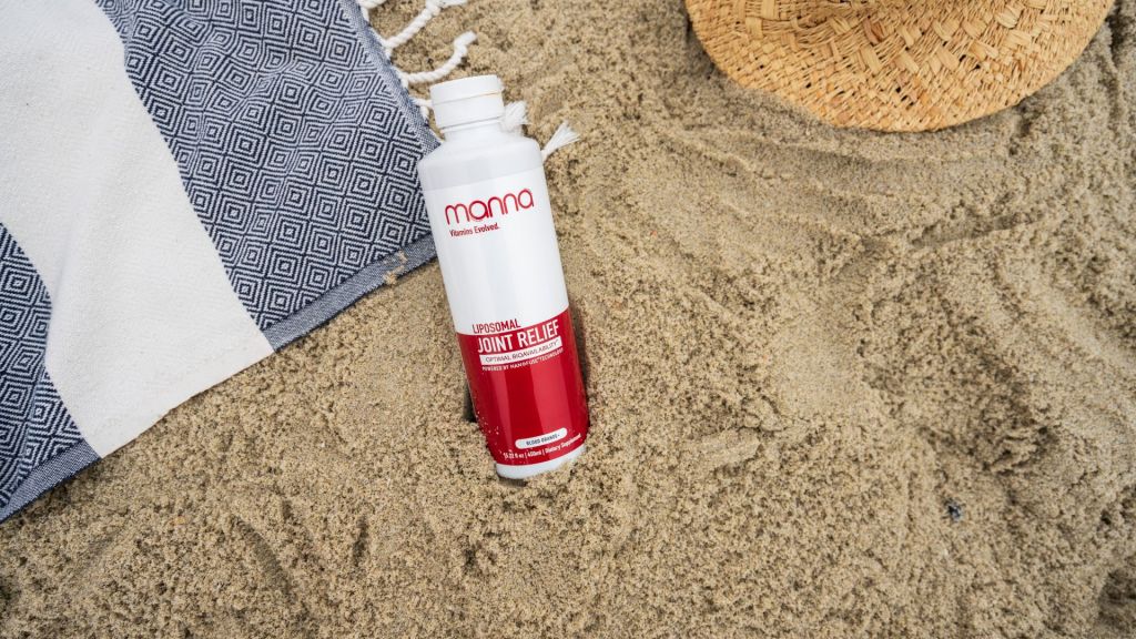 A bottle of liposomal joint relief cream lies on the beach sand next to a geometric-patterned towel and a straw hat.