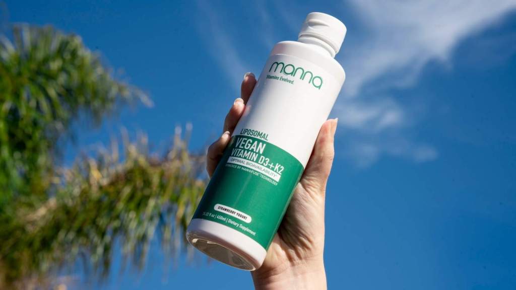 Hand holding a bottle of vegan vitamin D3+K2 supplement outdoors against a blue sky.