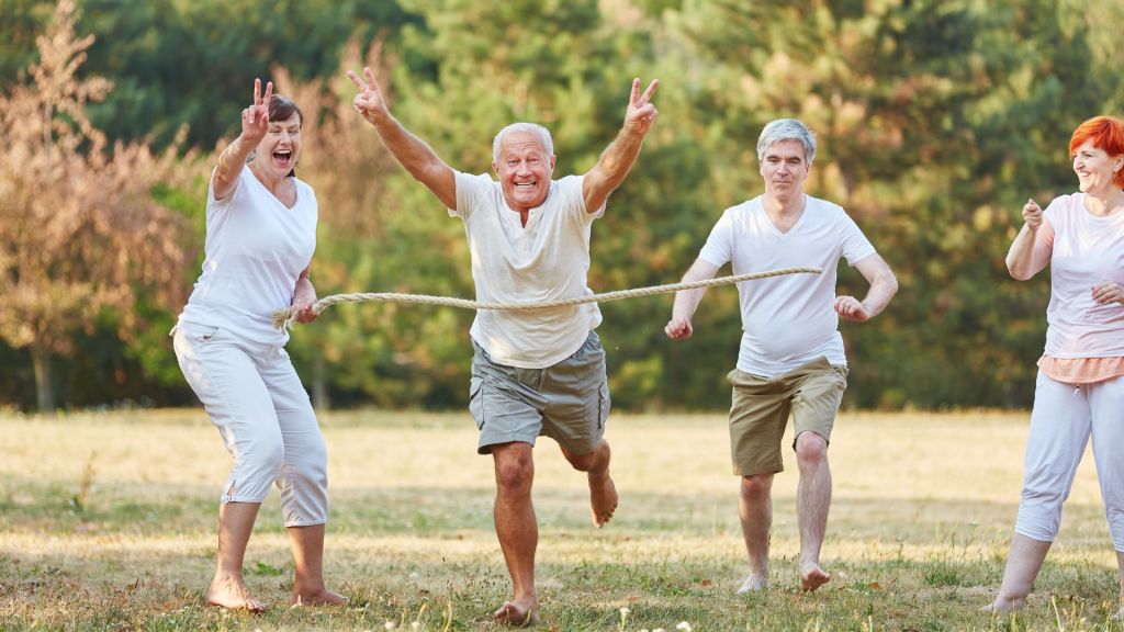 Four elderly people participate in a fun race outdoors. They are running barefoot and smiling while one man crosses the finish line holding up peace signs. Trees are visible in the background.