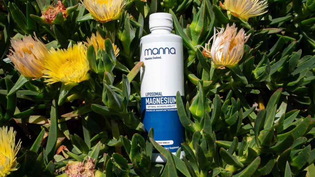 A white bottle of Manna Liposomal Magnesium sits upright among green leaves and yellow flowers outdoors.