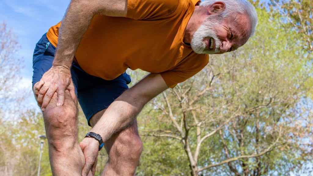 Older man outdoors in athletic clothing bends over, grimacing in pain and holding his knee, suggesting discomfort or injury. Trees and blue sky are visible in the background.